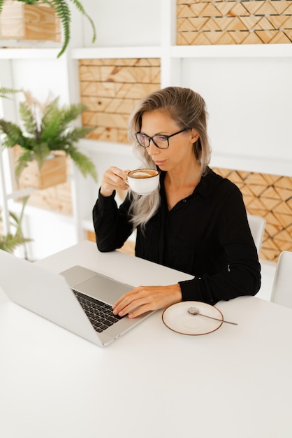 Geschäftsfrau im stilvollen Freizeitoutfit sitzt im Büro und benutzt Laptop und trinkt Kaffee