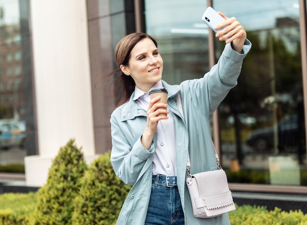 Geschäftsfrau im Trenchcoat macht Selfie am Telefon und trinkt Kaffee unterwegs im Stadtlebensstil