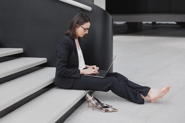 Geschäftsfrau in formellem Anzug mit Laptop im Büro sitzt im Stress von Terminen und komplexen Projekten auf den Stufen