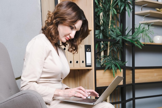 Geschäftsfrau in weißer Bluse sitzt auf einem Sessel in ihrem Büro mit einem Laptop auf dem Schoß