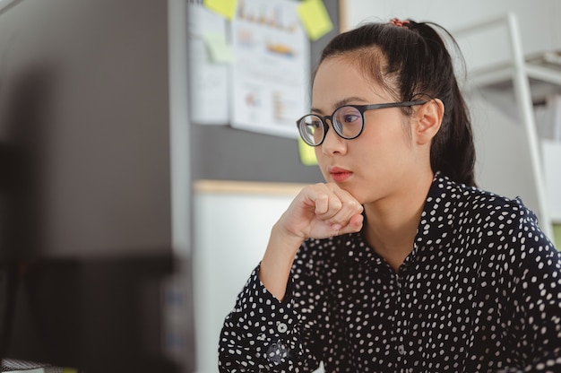 Geschäftsfrau mit Brille, die am Schreibtisch im Büro sitzt, das Kinn auf ihre Hände legt und wegschaut