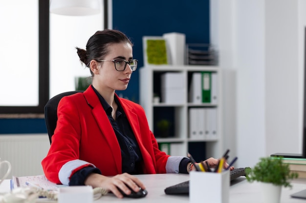 Geschäftsfrau mit Brille, die am Schreibtisch im Startup-Büro arbeitet. Selbstbewusster Manager in roter Jacke, der auf der Computertastatur tippt. Fokussierter Start-up-Besitzer am Arbeitsplatz mit Desktop-PC.