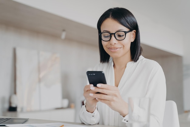 Geschäftsfrau mit Brille, die mobile Apps im Büro verwendet, liest gute Nachrichten, die online chatten