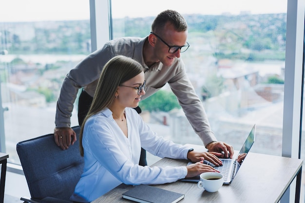 Geschäftsfrau mit einem laptop für einen männlichen kollegen, der in ein offenes notizbuch schaut geschäftliche zusammenarbeit und teamwork europäische millennials am tisch im büro moderne erfolgreiche menschen