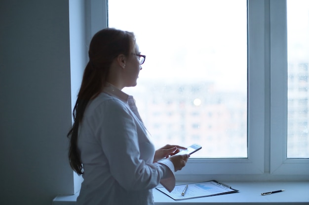 Geschäftsfrau mit einem Smartphone, während sie in der Nähe eines Bürofensters steht. Geschäftskonzept
