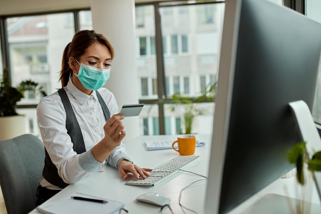 Geschäftsfrau mit Gesichtsmaske mit Kreditkarte, während sie ihr Online-Bankkonto auf einem Computer im Büro überprüft