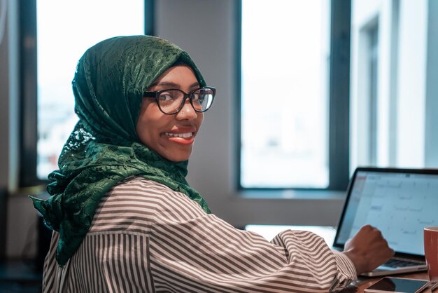 Geschäftsfrau mit grünem Hijab und Laptop im Ruhebereich bei modernem Großraum-Startup vor...