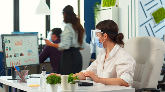 Geschäftsfrau mit Visier und Schutzmaske, die in einem neuen normalen Finanzgeschäftsbüro arbeitet. Mitarbeiter, die im Hintergrund beraten, das Unternehmensteam respektiert die soziale Distanz während der globalen Pandemie.