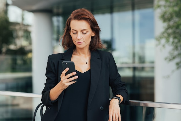Geschäftsfrau mittleren Alters in schwarzer Kleidung, die draußen auf ihrem Smartphone steht