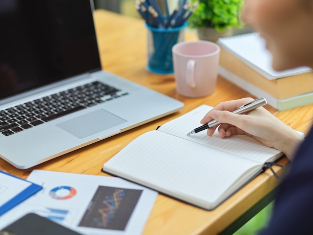 Geschäftsfrau Notizen über Notebook am Arbeitstisch mit Laptop, Finanzbericht, Kaffeetasse. Nahaufnahme