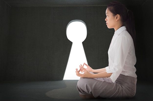 Geschäftsfrau sitzt in Lotus-Pose vor grauem Raum mit Schlüssellochtür