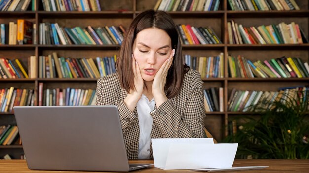 Geschäftsfrau tippt Antwort auf grauem Laptop mit Blick auf Bankkonto und legt verzweifelt den Kopf auf die Hände im Büro mit Bücherregalen