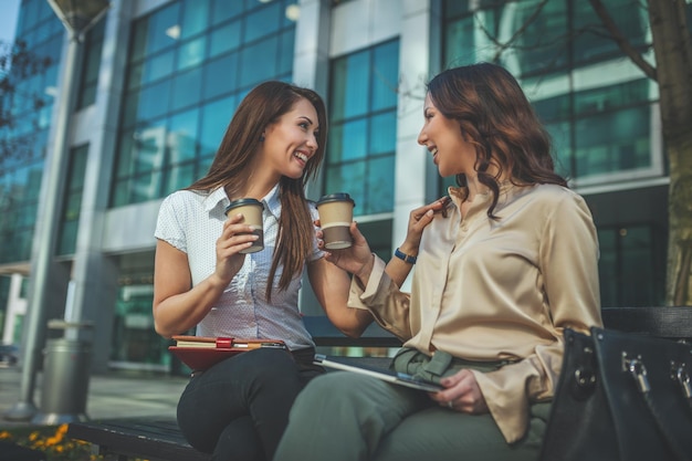 Geschäftsfrauen sitzen im Büropark, während sie ein digitales Tablet benutzen und eine Einweg-Kaffeetasse halten. Das Geschäftsteam arbeitet online zusammen, während es sich berät.