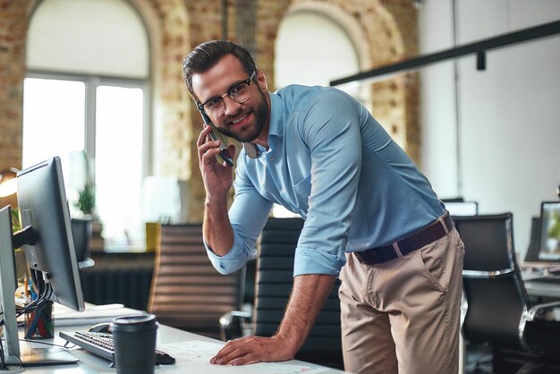 Geschäftsgesprächsporträt eines jungen und fröhlichen bärtigen Mannes mit Brille, der auf Smartphone spricht