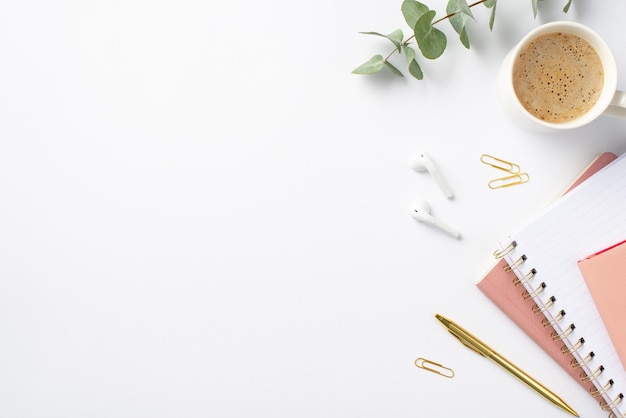 Foto geschäftskonzept draufsichtfoto eines arbeitsplatzstapels aus rosa planern, goldenen stiftklammern, eukalyptus, einer tasse kaffee und kabellosen ohrhörern auf isoliertem weißem hintergrund mit kopierraum