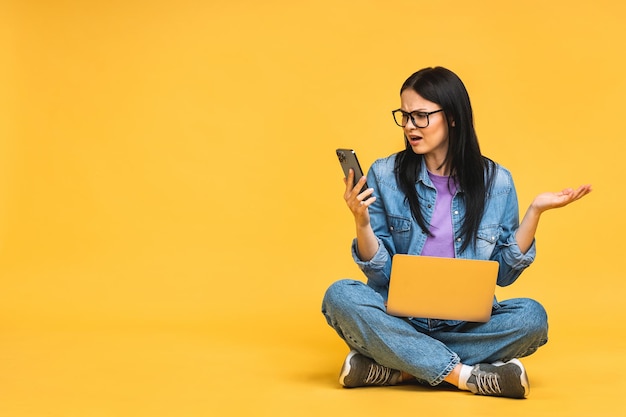 Geschäftskonzept Porträt einer depressiven, wütenden Frau in lässigem Sitzen auf dem Boden in Lotus-Pose und Halten eines Laptops isoliert über gelbem Hintergrund Mit Telefon