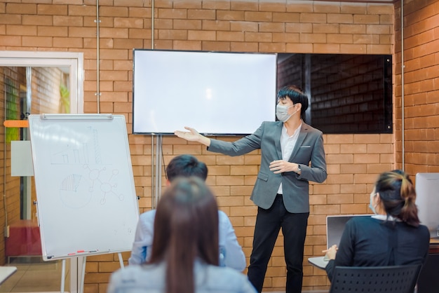 Geschäftskooperation: Junge asiatische männliche Trainer oder Sprecher machen Flipchart-Präsentation für verschiedene Geschäftsleute bei Treffen im Büro Männlicher Tutor oder Trainer stellt verschiedenen Kollegen das Projekt vor.