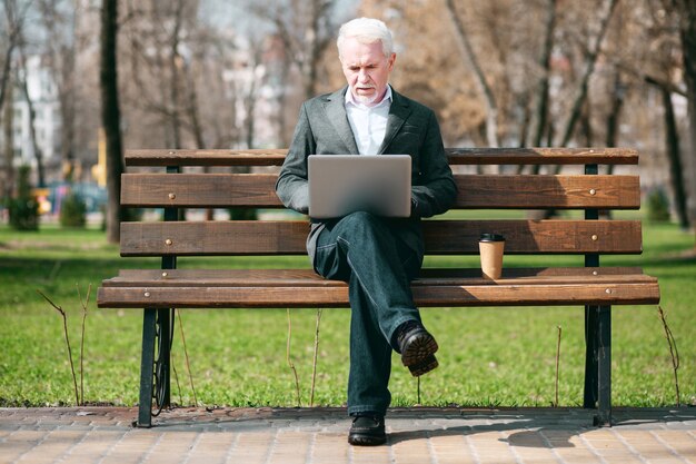 Geschäftskorrespondenz. Fokussierter reifer Geschäftsmann, der Laptop beim Sitzen im Park verwendet