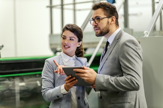 Geschäftsleute in einer Arbeitsumgebung in einer Fabrik. Ein Mann und eine Frau im eleganten Anzug begutachten die Arbeit in der Produktion. Sie halten ein Tablet in der Hand, während die Dame dem Mann den Vorgang erklärt