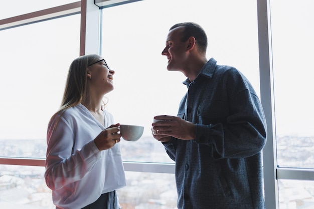 Geschäftsleute stehen entspannt in einer Arbeitspause bei einer Tasse Kaffee in einem modernen, von Glas umgebenen Büro mit Blick auf ein Hochhaus