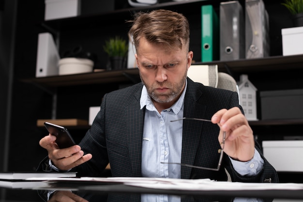 Geschäftsmann, der am Computertisch mit einem Telefon in seinen Händen sitzt und an Dokumente denkt