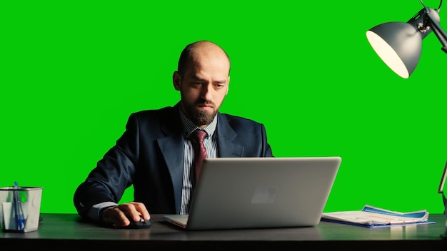 Geschäftsmann, der am Laptop vor Greenscreen-Hintergrund arbeitet und Dokumente am Schreibtisch verwendet. Erfolgreicher Teamleiter im Anzug posiert über isolierter Chroma-Key-Vorlage, leerer Copyspace-Hintergrund.