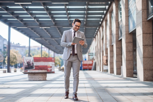 Geschäftsmann, der auf der Straße geht und Tablette benutzt, um eine E-Mail zu überprüfen. Im Hintergrund Business Center Exteriour.