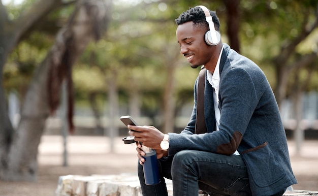 Geschäftsmann, der auf seinem Telefon in sozialen Medien surft und seine Musik-Playlist-App im Freien über Kopfhörer genießt Junger glücklicher und lässiger afrikanischer Mann, der sich während einer Pause in einem Naturpark draußen entspannt