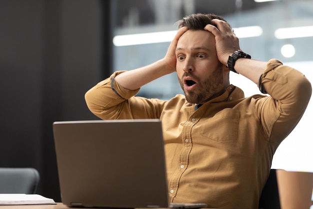 Geschäftsmann, der den Kopf berührt und im Büro unter Schock auf den Laptop schaut