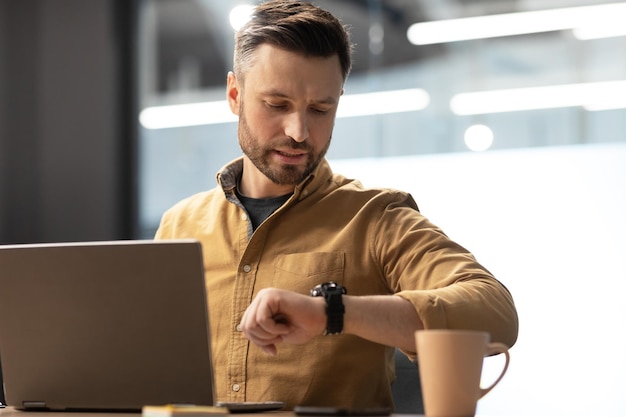 Geschäftsmann, der die Zeit mit Laptop überprüft und auf die Uhr im Büro schaut