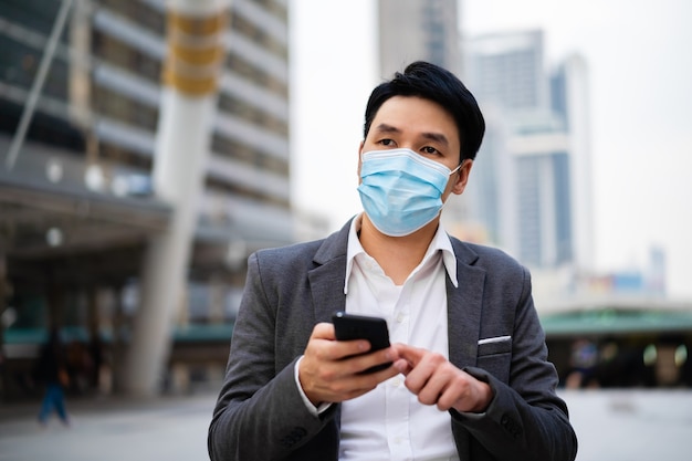 Geschäftsmann, der ein Smartphone verwendet und medizinische Maske während der Coronavirus-Pandemie in der Stadt trägt