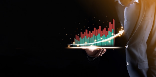 Geschäftsmann, der ein wachsendes virtuelles Hologramm von Statistik, Grafik und Diagramm mit Pfeil oben auf dunklem Hintergrund hält und zeigt. Aktienmarkt. Geschäftswachstum, Planung und Strategiekonzept.