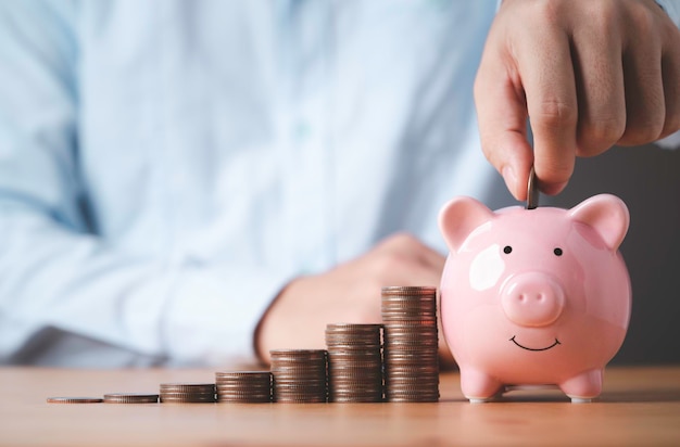 Geschäftsmann, der eine Münze in ein rosa Sparschwein legt, wobei sich die Münzen auf dem Tisch stapeln Geld sparen und Einzahlungen für Investitionen, um ein Gewinn- und Dividendenkonzept zu erhalten