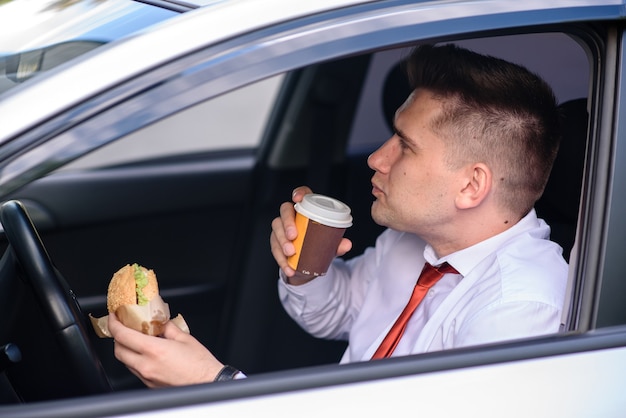 Geschäftsmann, der einen Burger isst und Kaffee im Auto trinkt.