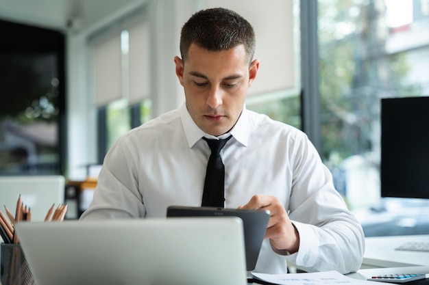 Geschäftsmann, der im Büro mit Tablet-Laptop und Dokumenten auf seinem Schreibtisch arbeitet und Daten analysiert