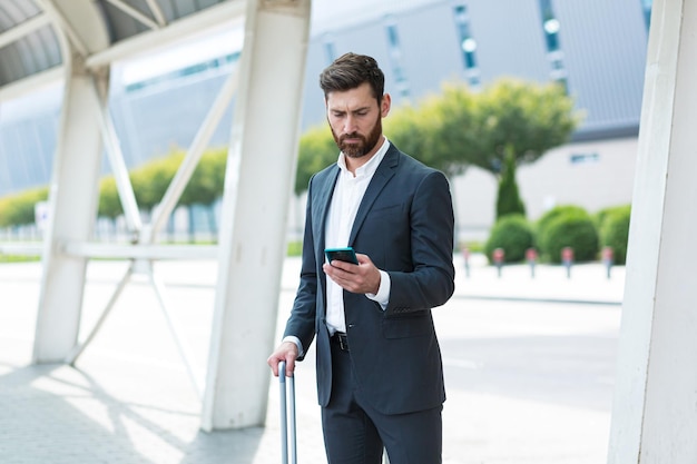 Geschäftsmann, der im Hintergrund steht, ein moderner Bahnhof Flughafen oder eine Bushaltestelle in formellem Anzug mit einem Koffer mit App-Handy Reisender Geschäftsmann Tourist mit einem Smartphone in der Hand bestellt ein Taxi