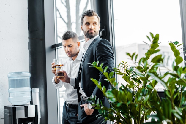 Geschäftsmann, der mit der Hand in der Tasche steht und ein Glas Whiskey in der Nähe eines Kollegen hält