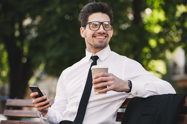 Geschäftsmann draußen im Park unter Verwendung des Mobiltelefons, das Kaffee trinkt.