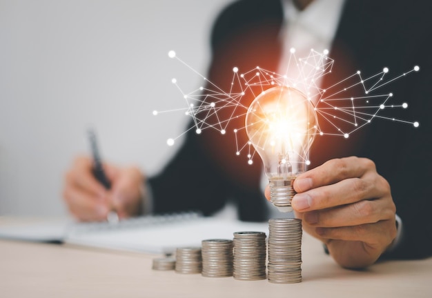 Geschäftsmann hält Glühbirne mit Münzen, die auf dem Schreibtisch gestapelt sind, um Energie und Geld zu sparen die Idee mit Innovations- und Inspirationskonzept, das Energie und Geld spart