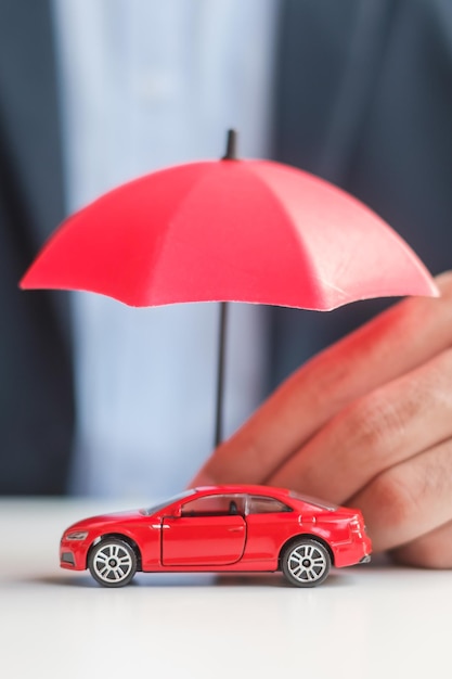 Geschäftsmann Hand mit Regenschirm und Deckel rotes Auto Spielzeug auf dem Tisch Reparatur der Autoversicherung Garantie Finanzielles Banking und Geldkonzept