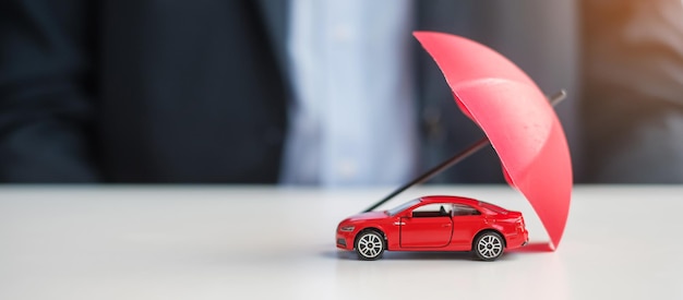 Geschäftsmann Hand mit Regenschirm und Deckel rotes Auto Spielzeug auf dem Tisch Reparatur der Autoversicherung Garantie Finanzielles Banking und Geldkonzept