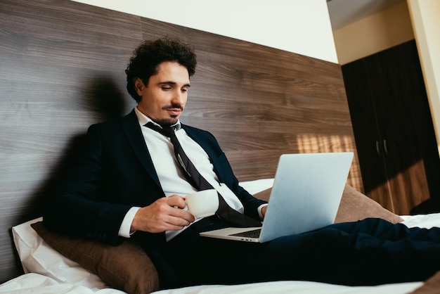 Geschäftsmann im Anzug hält eine Tasse Kaffee und arbeitet am Laptop im Hotelzimmer