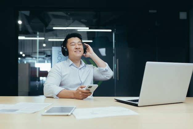 Geschäftsmann im Büro ruht sich während der Arbeitspause aus und hört Musik in großen schwarzen Kopfhörern asiatischer Mann