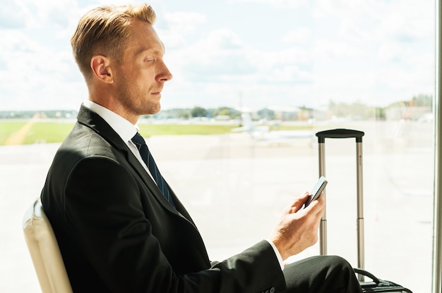Geschäftsmann im Flughafen. Seitenansicht eines selbstbewussten Geschäftsmannes in formeller Kleidung, der ein Mobiltelefon hält, während er auf einen Flug am Flughafen wartet