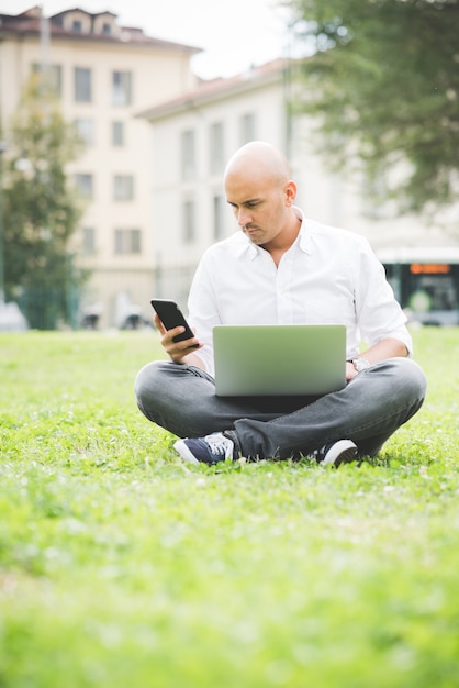 Geschäftsmann im weißen Hemd, das mit dem Laptop sitzt auf dem Gras in einem Park arbeitet