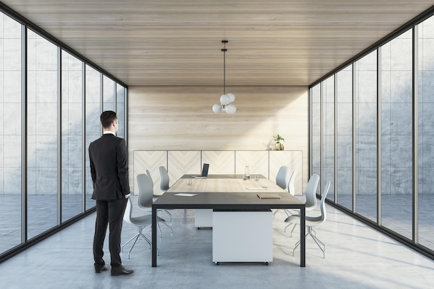 Geschäftsmann in Anzug, der in einem modernen Innenraum aus Holz und Beton mit Panorama-Glasfenstern und Tisch mit Geräten steht