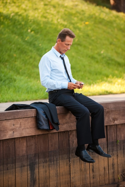 Geschäftsmann in der Natur. Zuversichtlich reifer Geschäftsmann, der Handy hält und es beim Sitzen am Kai anschaut