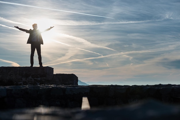 Geschäftsmann in einem Anzug, der bei Sonnenuntergang von einem Sonnendurchbruch beleuchtet wird, während er mit ausgestreckten Armen an einer Wand steht, die vom Licht mit Kopierraum in einem konzeptionellen Bild silhouettiert wird