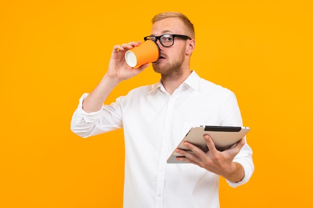 Geschäftsmann in einem weißen Hemd mit einer Tablette und einer Tasse Kaffee in der Hand auf einem gelben Hintergrund mit Kopienraum.