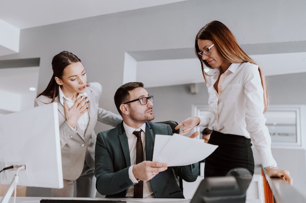 Geschäftsmann in formeller Kleidung, der mit seinen Kolleginnen über Dokumente spricht, während er im modernen Büro sitzt.